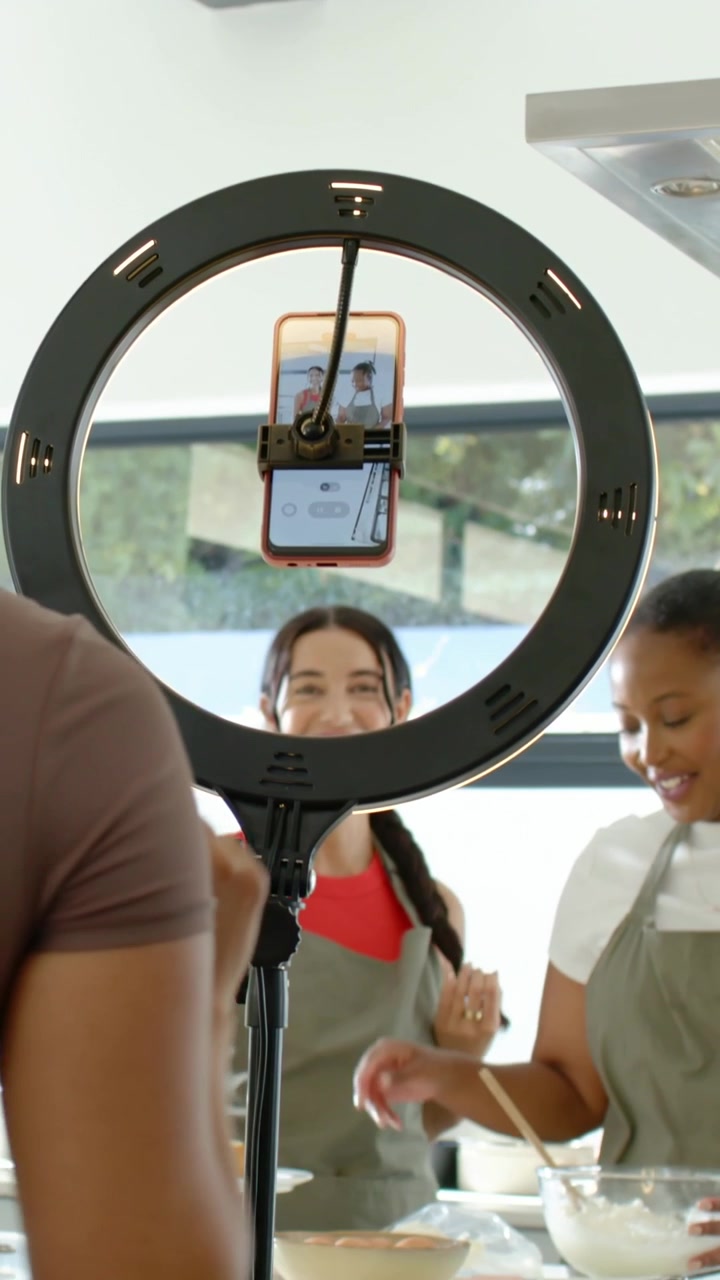 Vertical video: Tapping braided host framing phone in LED light in kitchen, cohost in apron mixing