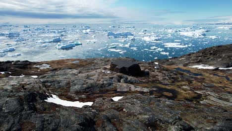 Remote-scientist-station-on-rocky-shore-of-Greenland-with-icebergs-in-background,-aerial-view