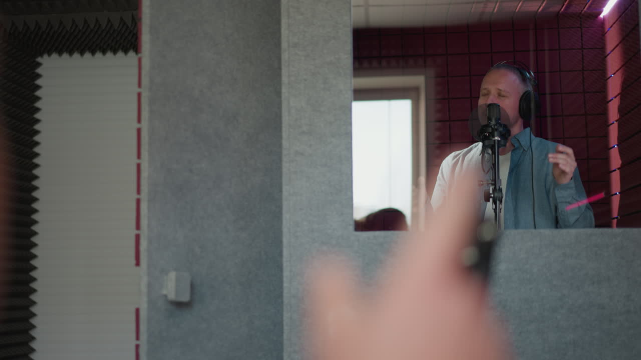 한 남자가 검은 헤드폰과 파란색 장갑 셔츠를 입고 녹음 스튜디오에서 열정적으로 노래합니다. 그가 공연하는 동안 그의 눈은 닫혀 있고, 손의 몸짓은 거울을 통해 포착됩니다.