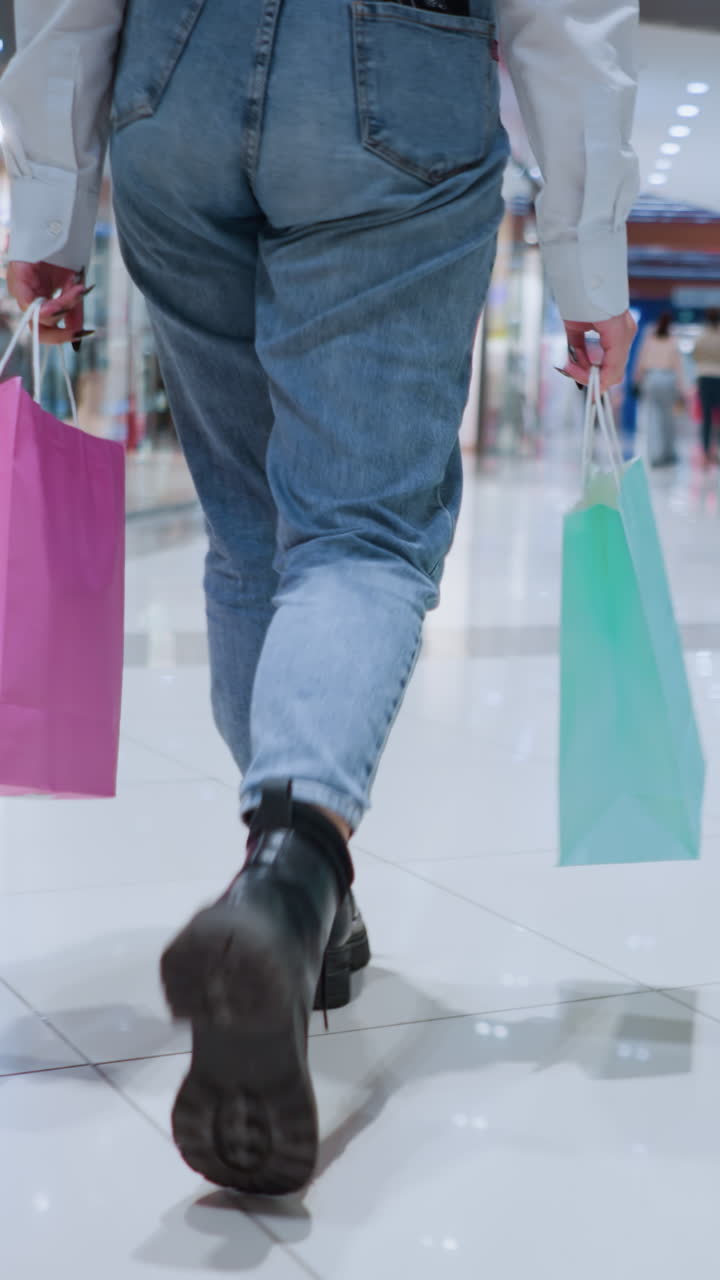 안경을 쓴 자신감 넘치는 여성이 양손에 다채로운 쇼핑백을 들고 현대 쇼핑몰을 고 있습니다.