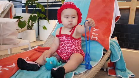 cute baby girl holding blue strap and water tumbler on lounge chair by the poolside