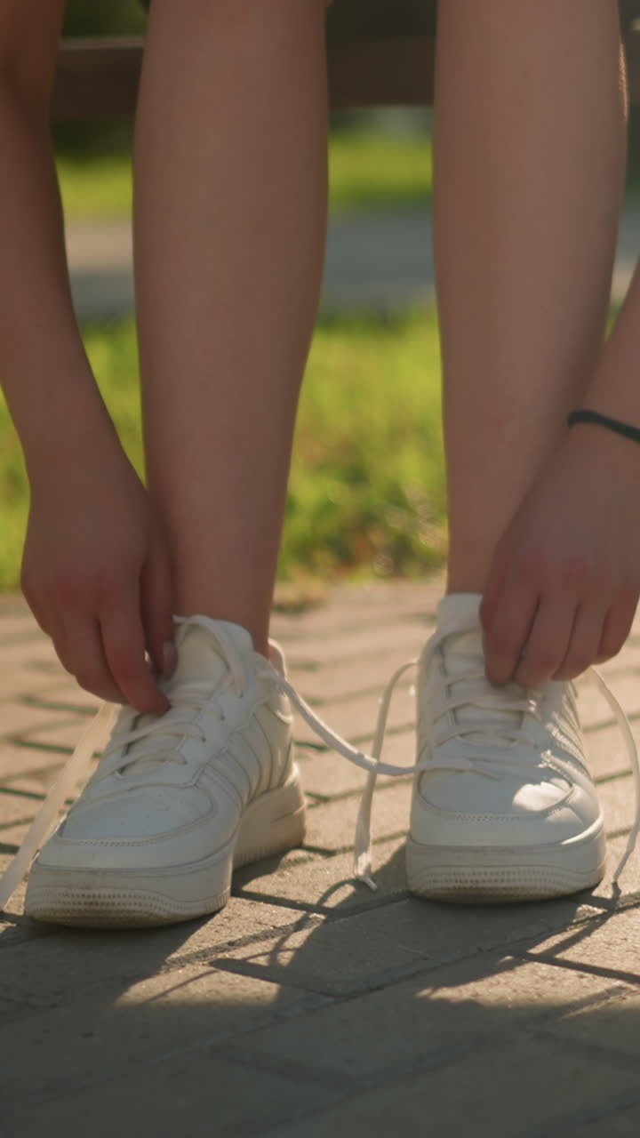 은 빛으로 벤치에 앉아 있는 여자가 스니커의 신발을 풀고 있는 모습. 뒷면에 초록색이 있는 보행로에 부드러운 그림자를 던지고 있다.