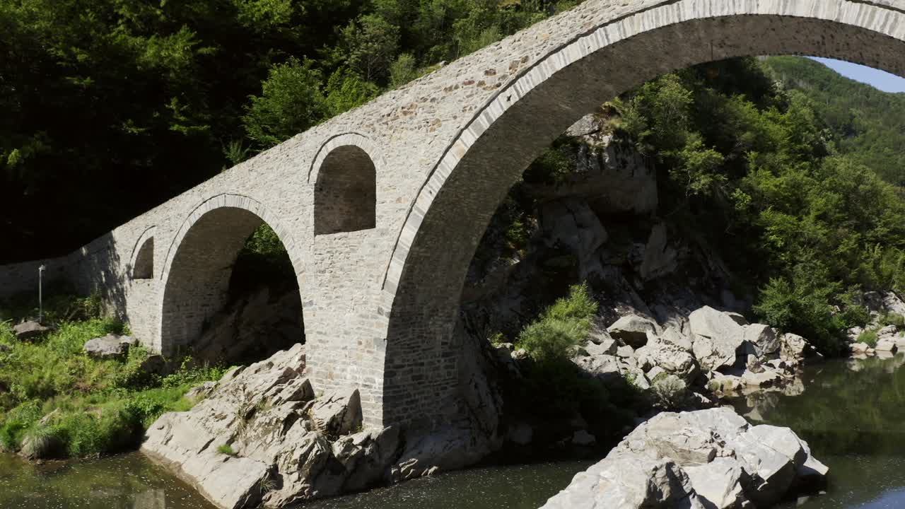Retreating drone shot of the arches of the Devil's Bridge, and panning to reveal the Arda River, located in the town of Ardino in Bulgaria