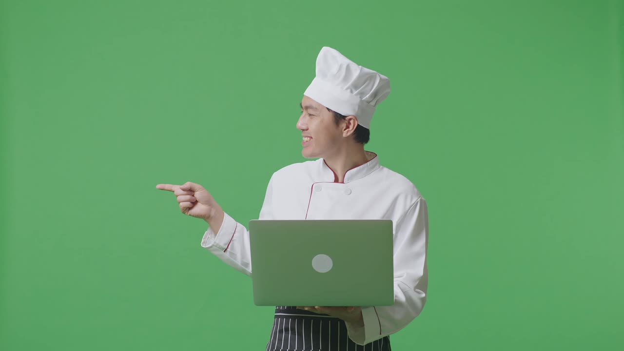 Asian Man Chef Using A Laptop And Pointing To Side While Standing In The Green Screen Background Studio