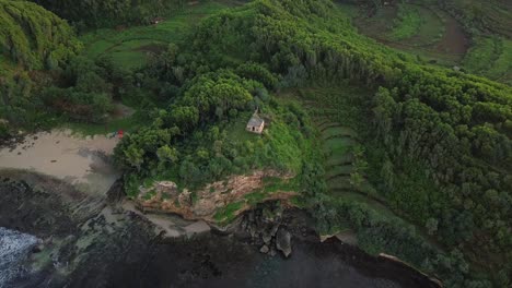 toma aérea de arriba hacia abajo que muestra un edificio antiguo en la cima de una colina con un acantilado, un océano y una playa de arena privada.
