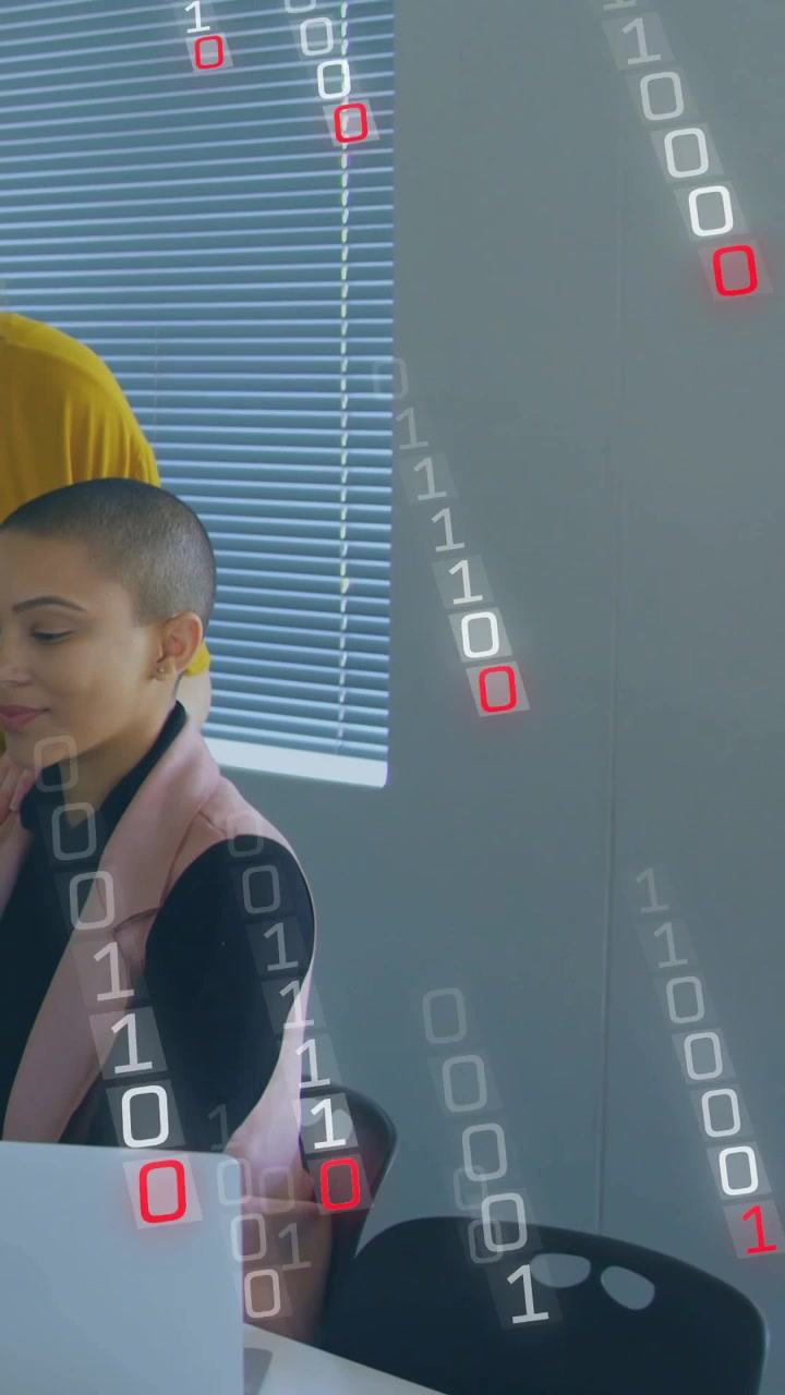 Vertical video: Woman using laptop with IT peer watching, binary falling onto laptop for review