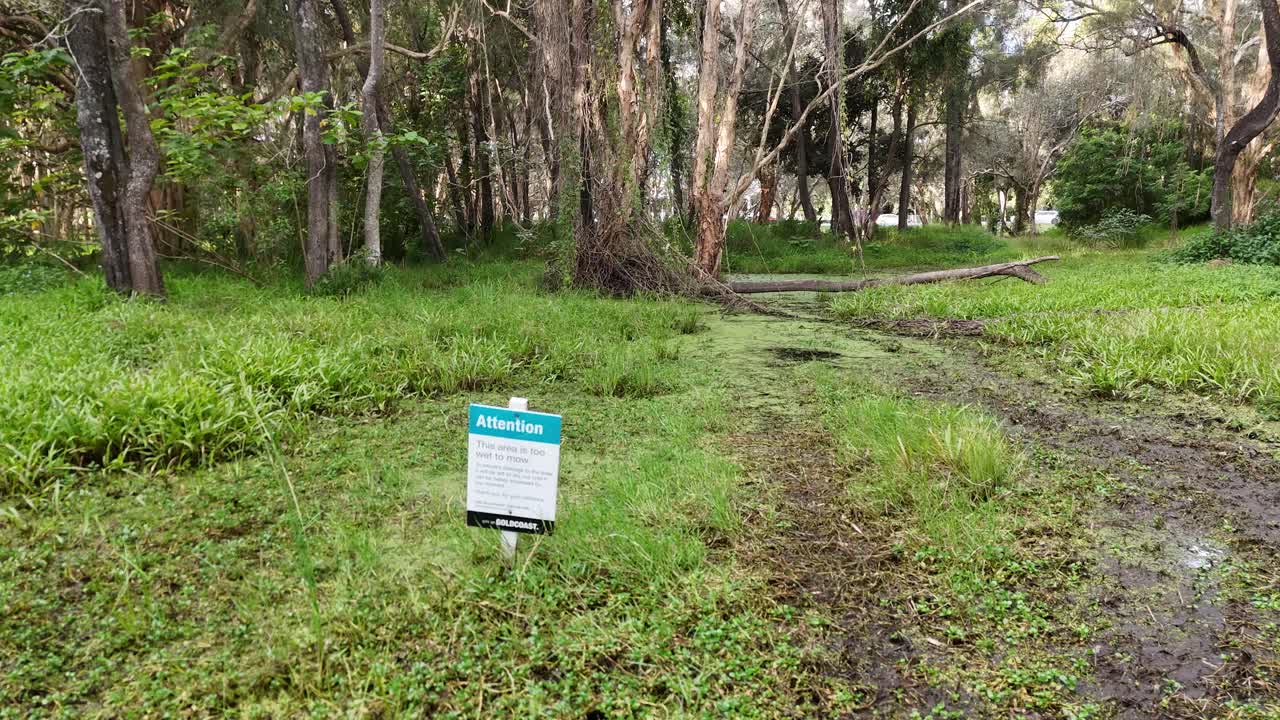 Drone captures a wet, empty park with warning sign amidst lush greenery and eucalyptus trees on a rainy day