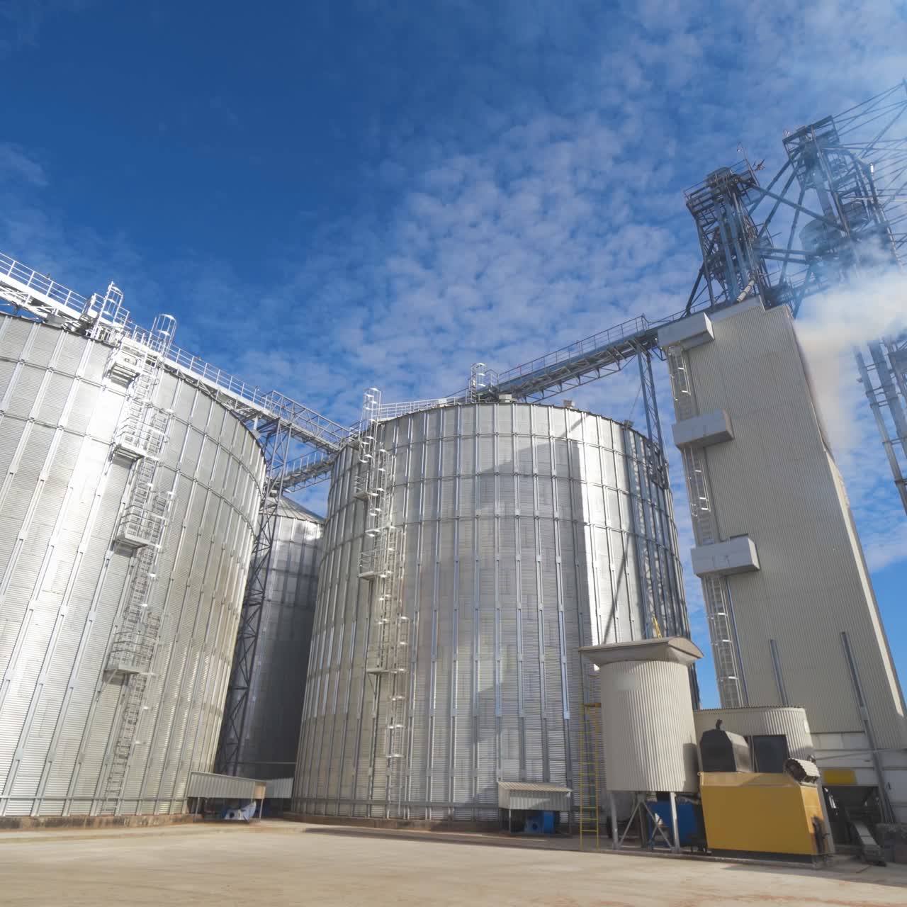 Huge hangar buildings. Modern grain elevators under blue sky. White vapor released from metal construction for drying grains. Panoramic view