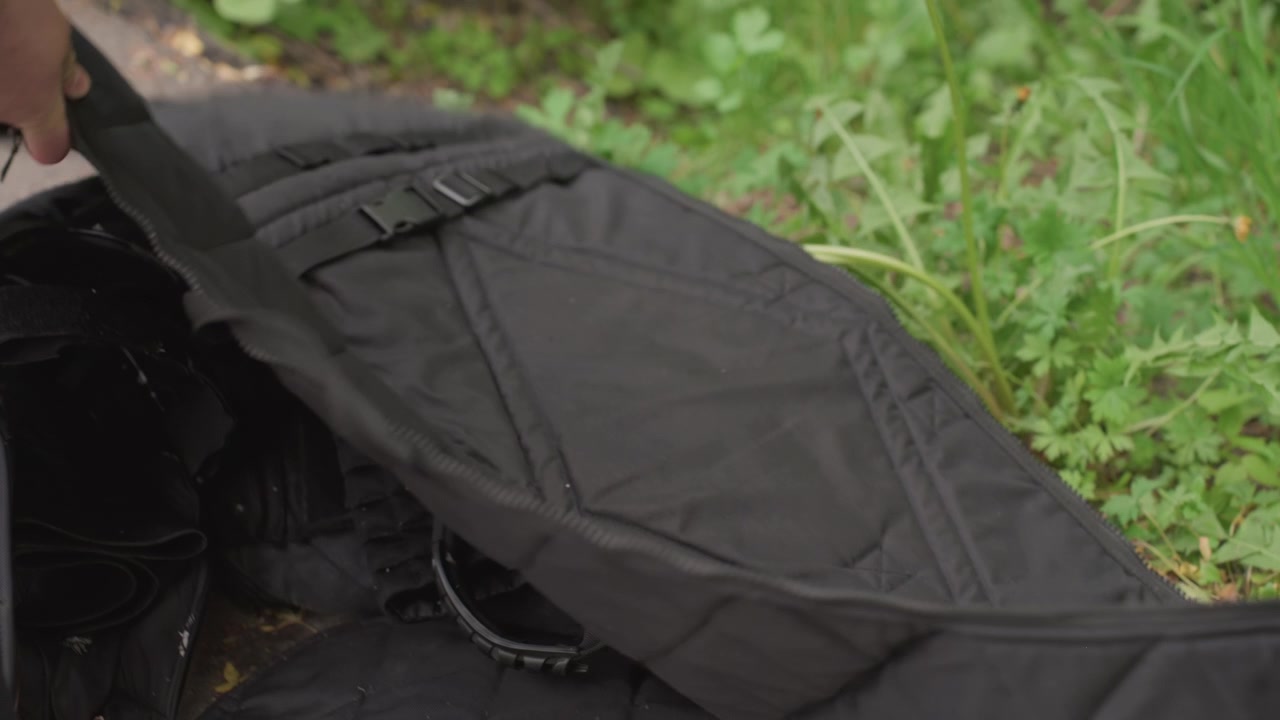 屋外用の黒いパッド入りバッグのジッパーを開け、ストラップを調整している人物の手のクローズアップ。バッグの内部コンパートメントが見えている。背景には緑の葉があり、自然環境を示している。