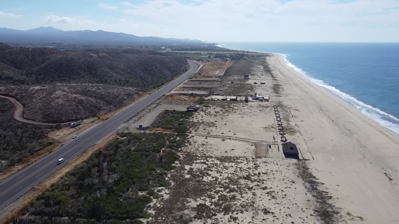 dolly aéreo filmado frente a la pintoresca costa de cabo san lucas en todos santos a lo largo de una carretera costera hacia baja california sur en méxico con una vista de la playa, coches en movimiento y el mar azul