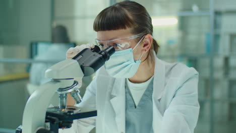 female lab worker in mask working with microscope
