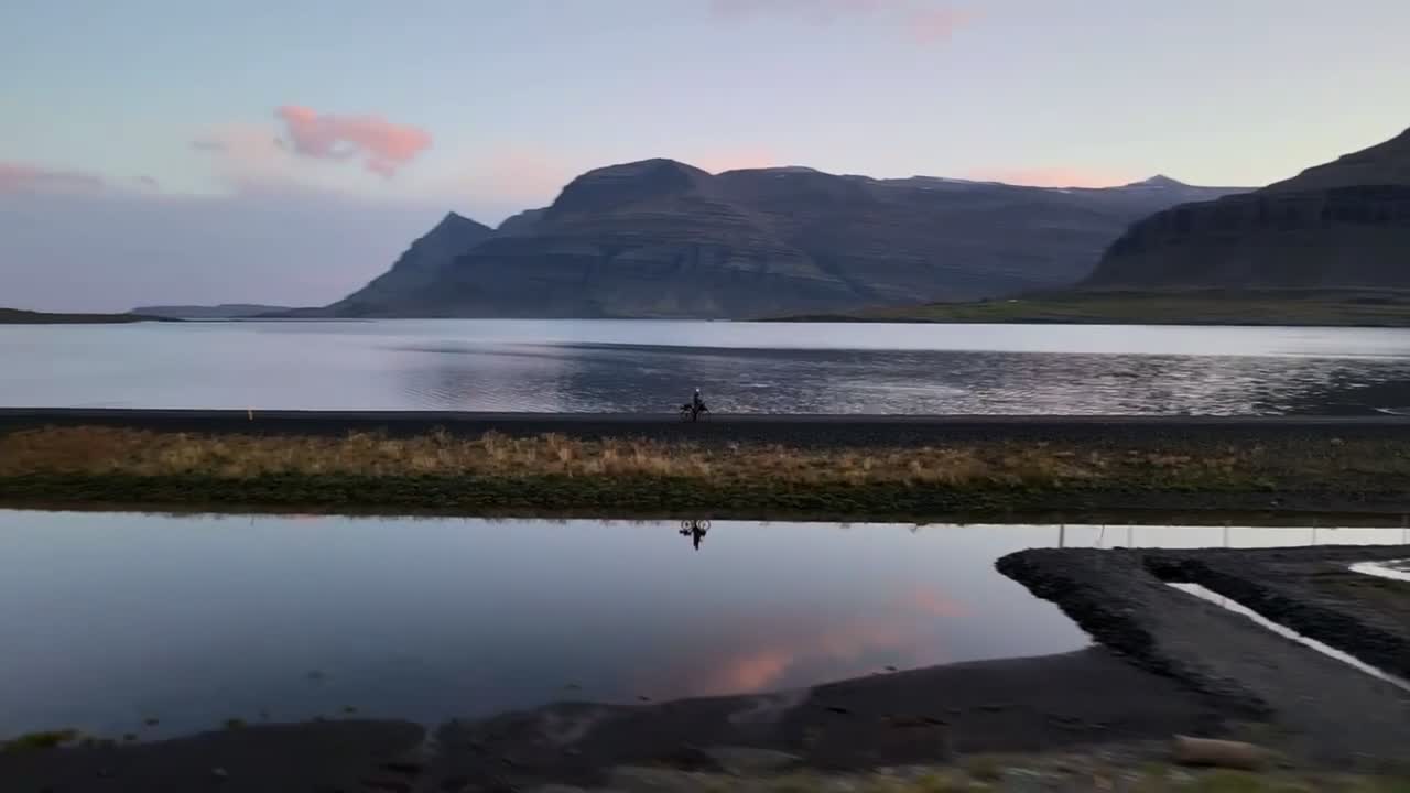 Aerial Spiral Away from Bicycle on Reflective Iceland Lake with Mountains