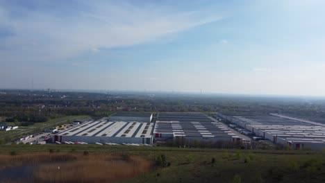 Edificio-Industrial-Logístico-Masivo-Con-Muchos-Camiones-Y-Vehículos,-Vista-Aérea-Desde-La-Cima-De-La-Colina