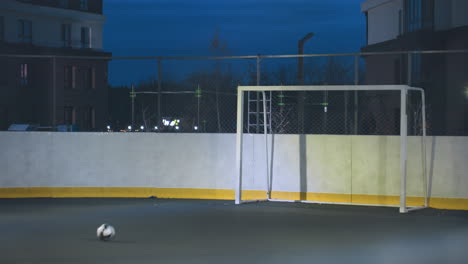 soccer ball enters goal post with precision and rolls out on illuminated outdoor field during night practice, surrounded by urban scenery with tall residential buildings and vibrant city lights