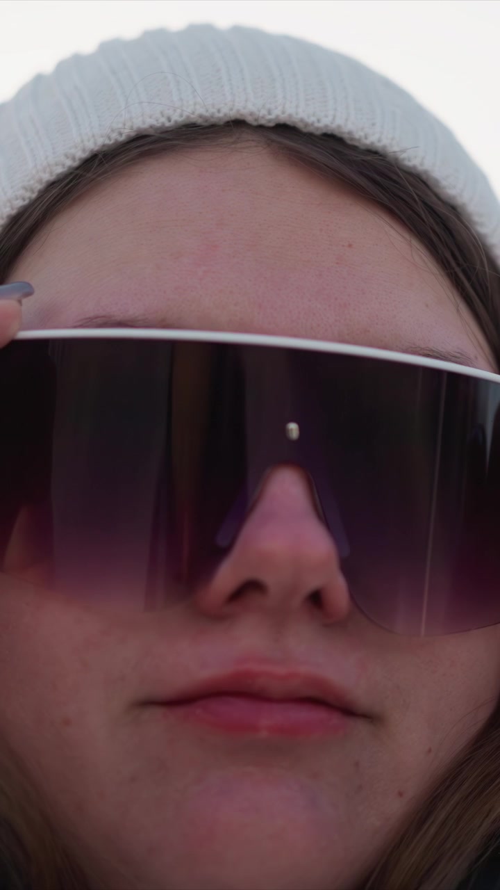 Primo piano di una donna con occhiali da sole che li aggiusta, con una luce naturale soffusa che ne esalta i tratti del viso. I capelli della soggetta sono visibili sotto un berretto di lana bianco, evidenziando i dettagli della texture della pelle