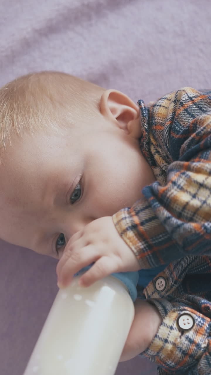 funny blond boy eats delicious food from plastic bottle with teat lying on soft bed with pink plaid in room close view