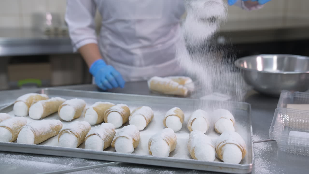 Savory cannoli sweets laid on the metal tray for decoration. Worker covers desserts with sugar powder from a sieve. Close up.