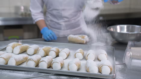 savory cannoli sweets laid on the metal tray for decoration. worker covers desserts with sugar powder from a sieve. close up.
