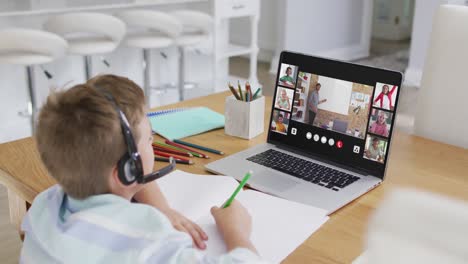 Schoolboy-using-laptop-for-online-lesson-at-home,-with-diverse-teacher-and-class-on-screen