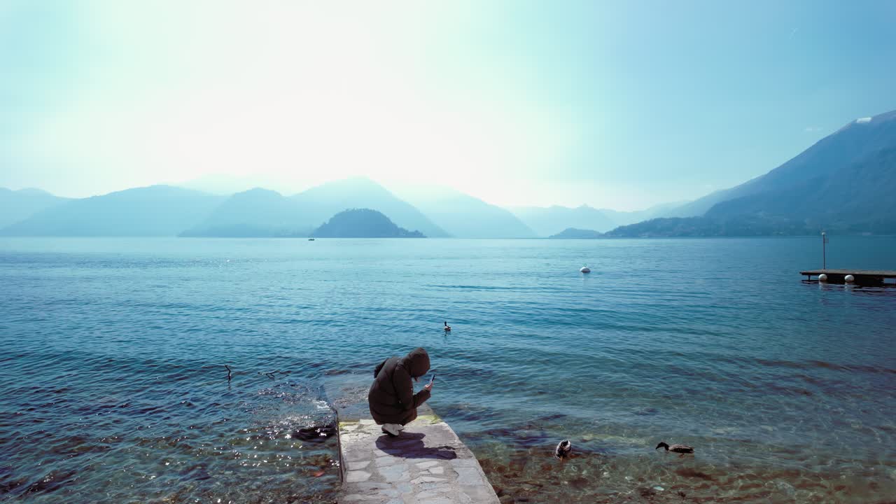 Woman at a pier filming the ducks near the shore, Varenna, Lake Como, Italy