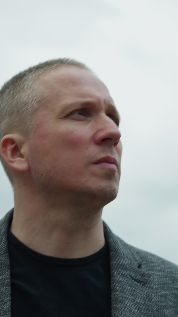 una vista cercana de un hombre blanco envejecido con un traje gris y una camiseta polo negra, volviéndose hacia atrás con una expresión enfocada, con un cielo nublado a la vista