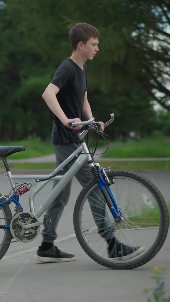 회색 바지와 검은색 을 입은 소년은 보행자 교차로를 가로질러 자전거를 고, 다른 자전거 타는 사람들이 배경에 보이고, 나무와 함께 몇 개의 표지판과 움직이는 자동차가 주변에서 볼 수 있습니다.