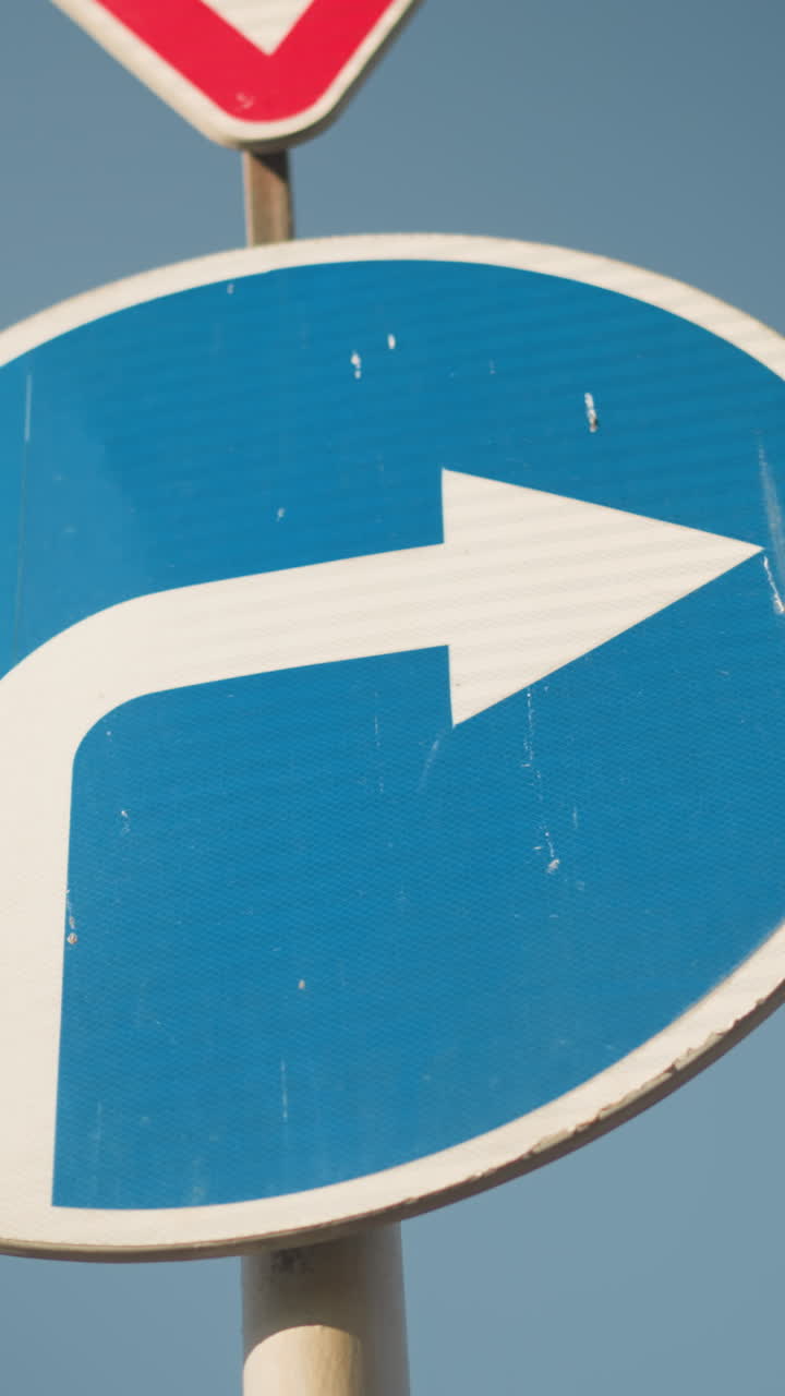 Blåt højresvingsskilt mod himmel, nærbillede af cirkulært skilt med hvid pil og trekant for ubetinget vigepligt over, falmet maling og tekstureret overflade, stangmontering under klart dagslys, formidler navigation