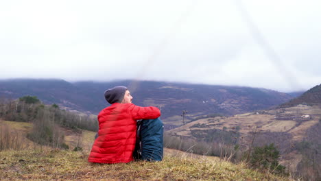 Young-hiker-sitting-in-green-natural-landscape-resting-with-his-modern-backpack-after-long-day-of-trekking-up-to-mountains-trails-discovering-unexplored-land