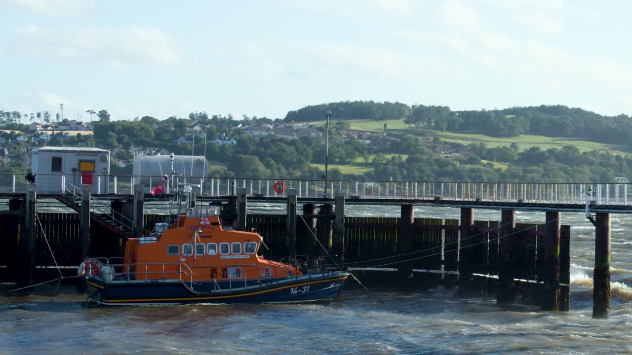 An orange lifeboat approaches and docks at a pier on a windy Scottish coastal day