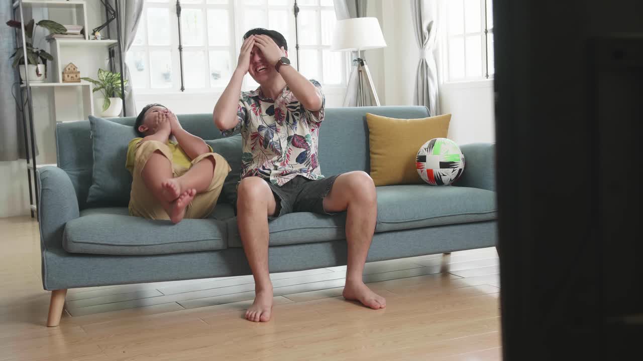 Padre asiatico frustrato e deluso e suo figlio stanno guardando la TV mentre sono seduti su un divano a casa.
