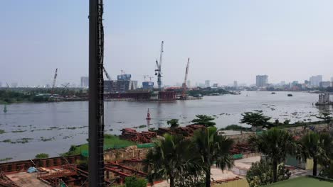 ascending-aerial-shot-of-ultra-modern-mobile-phone-tower-disguised-as-a-palm-tree-against-a-background-of-a-riverfront-construction-site-with-city-buildings,-cranes-and-boat-traffic-on-the-river