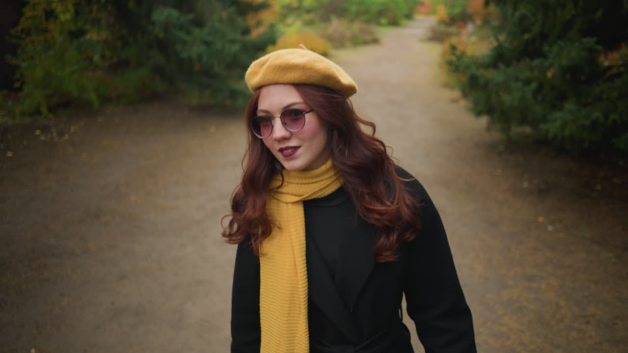Elegante signora con un basco giallo e una sciarpa inclina la testa a sinistra e a destra, osservando attentamente l'ambiente circostante mentre passeggia in un giardino vivace.