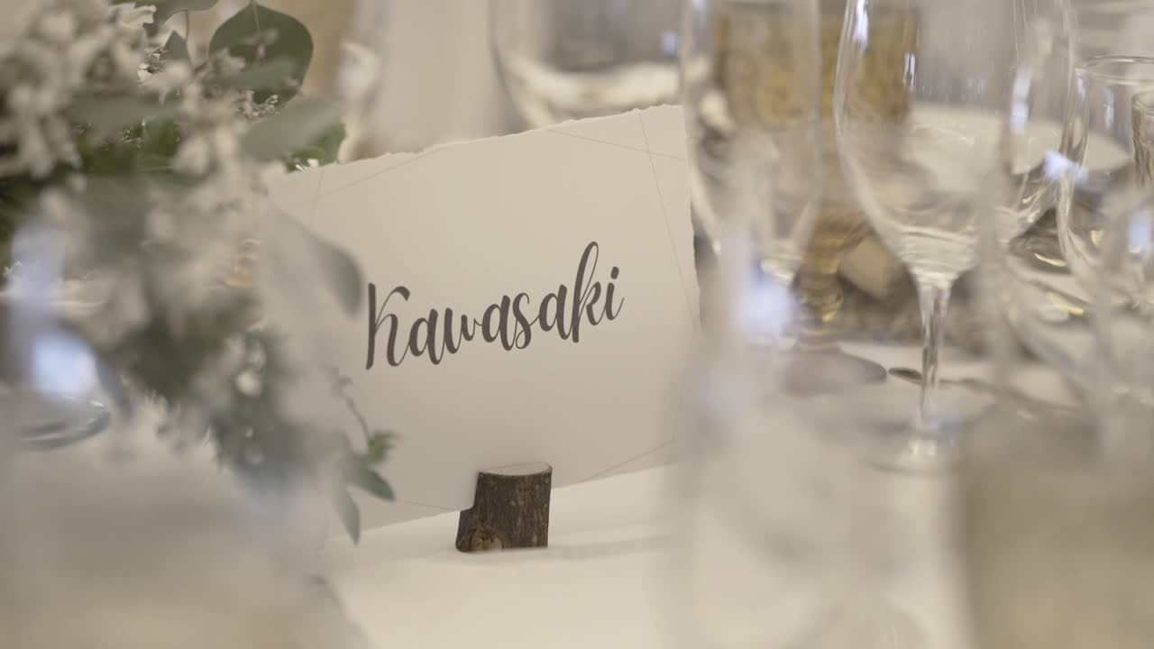 Close up of a wedding table card reading Kawasaki among glassware and floral decor