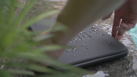 a black, basic, waterproof laptop case with lines engraved in it lies on the side of a pool with splashes and drops of water on it