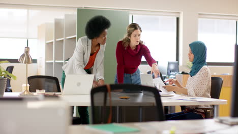 diverse female creative colleagues in discussion using laptop and notes in office, slow motion