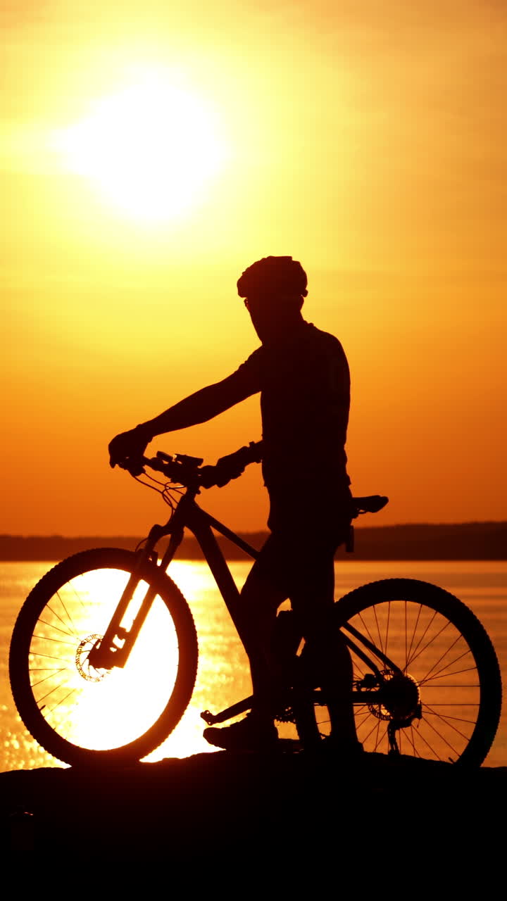 Silhouette of a male cyclist with helmet at sunset near the river. Bike rider walks with his bike on the background of the golden path from the sun over the water surface. Vertical video