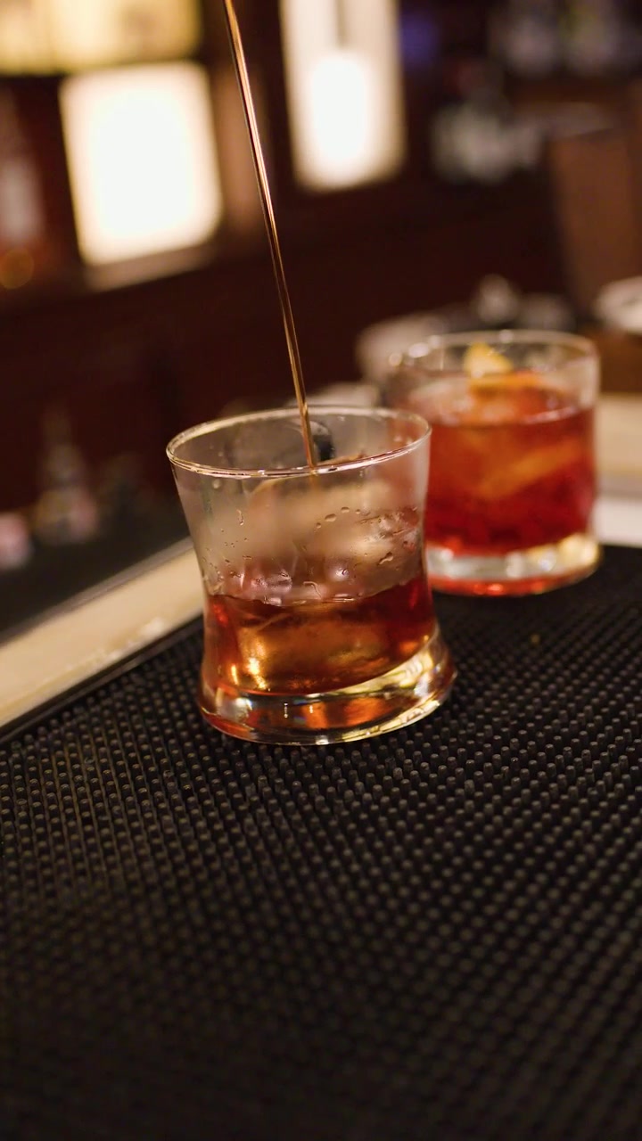 Un barista prepara e versa cocktail al whisky con ghiaccio in un ambiente da bar elegante e in penombra.