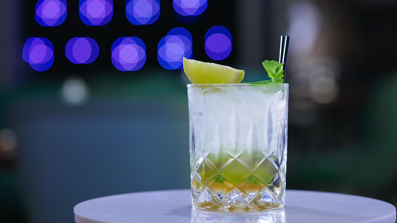 Misted glass full of beverage with two short black straws in it. Rotating glass with lemon and mint leaves on top. Close up. Blurred backdrop.