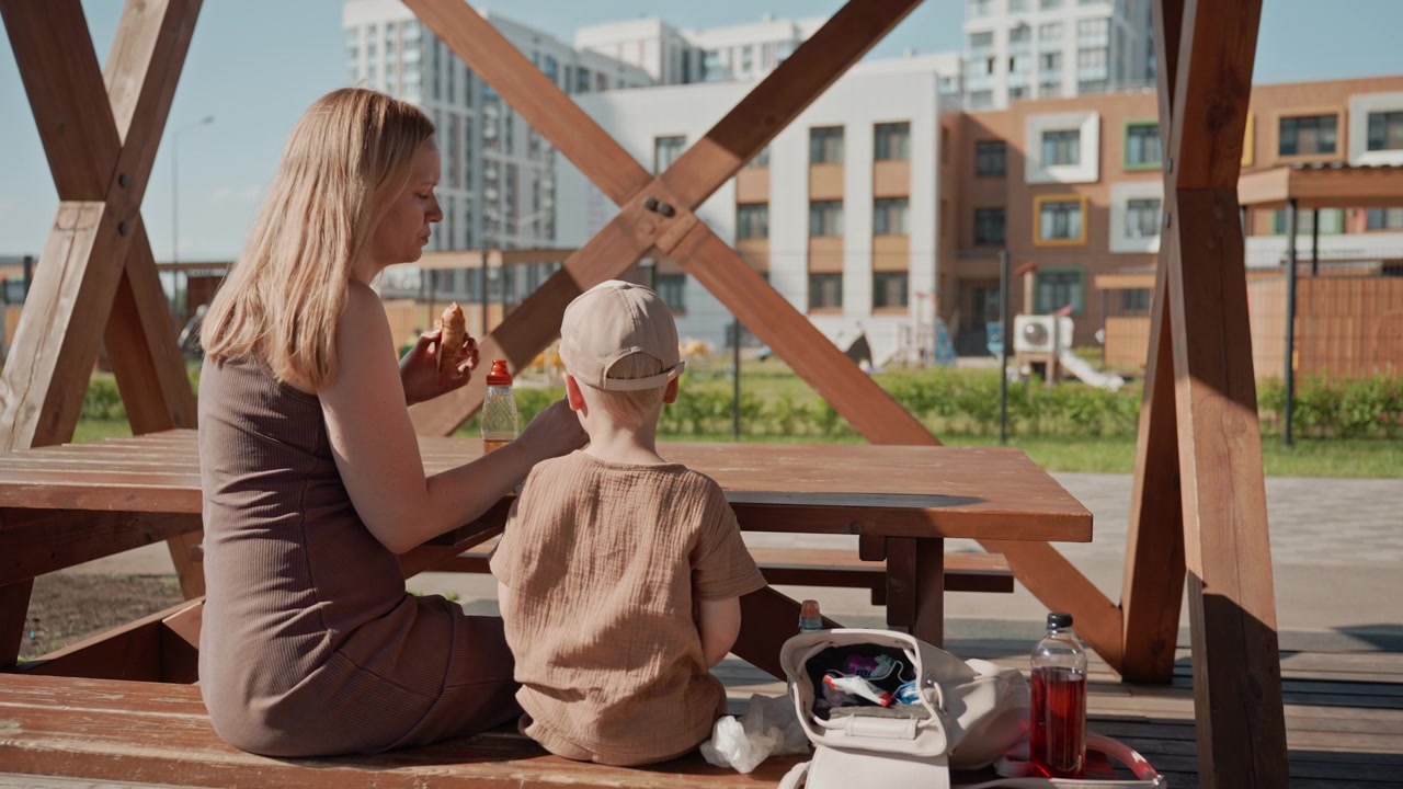 Kvinde og ung dreng sidder sammen ved et træpicnicbord udendørs og deler mad. Scenen inkluderer personlige genstande som flasker i en taske på bænken med moderne bygninger synlige i baggrunden.