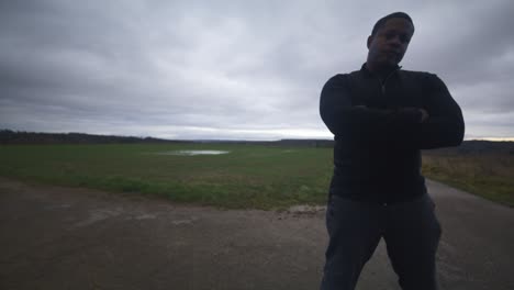 hombre negro moderno esperando en un campo abierto bajo un cielo gris, europa, 4k | musculoso, intimidante, sombra, figura, borroso, ropa oscura, poderoso, cercano, matón, bloqueo, obstáculo, malo, enojado, brazos cruzados