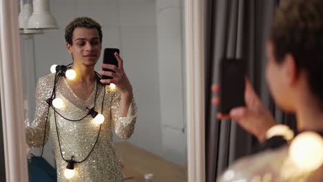 a man in a dress posing in front the mirror with garland on neck and glass of wine using phone