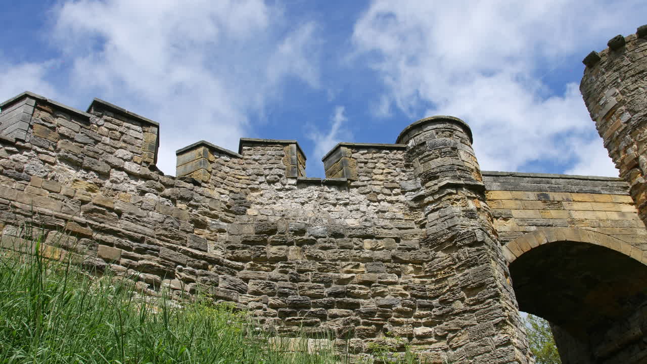 Stone castle walls with battlements and towers rise in Scarborough, North Yorkshire, England, viewed from below against blue sky and white clouds