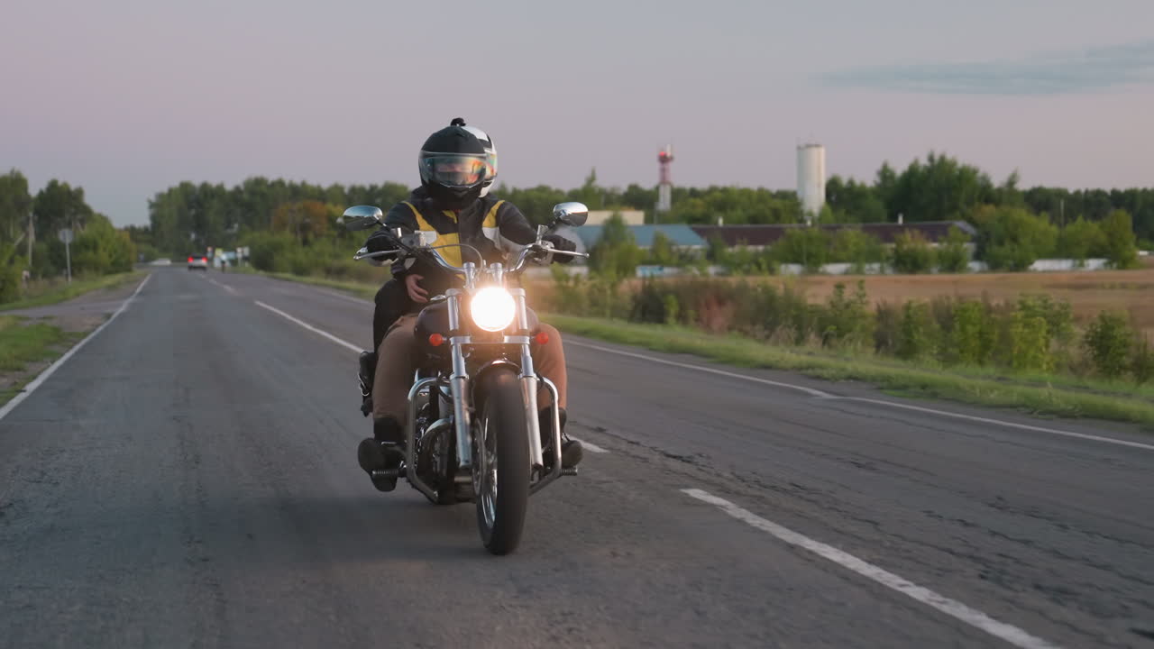 Motociclista con casco percorre una strada di campagna, con il faro cromato della moto che brilla mentre sullo sfondo si intravedono edifici industriali e una torre