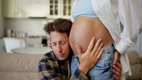 a middle-aged man in a plaid shirt puts his ear to the belly of his pregnant wife and listens to a small child during pregnancy at home