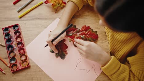 unknown girl circling oak leaf, laying on white sheet of paper, with black felt-tip pen. sitting at table at home. close up, top down view