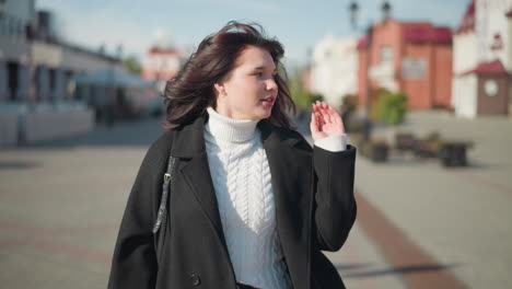 young woman adjusting her hair looking to her left, dressed in a white cable knit sweater and black coat, on a sunny urban street, exuding casual elegance