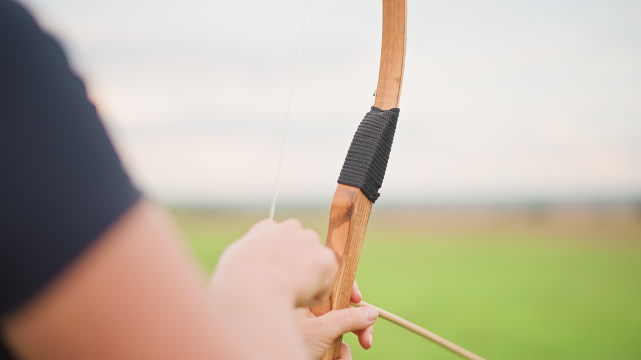 Kaukasischer Bogenschütze, der in einem grasbewachsenen Feld einen Holzbogen spannt, Nahaufnahmen des mit Leder umwickelten Griffs, Kundschafterhaltung wechselt zu Jäger und Krieger, Auszubildender konzentriert sich beim Zielen und Abschießen des Pfeils auf die Ferne