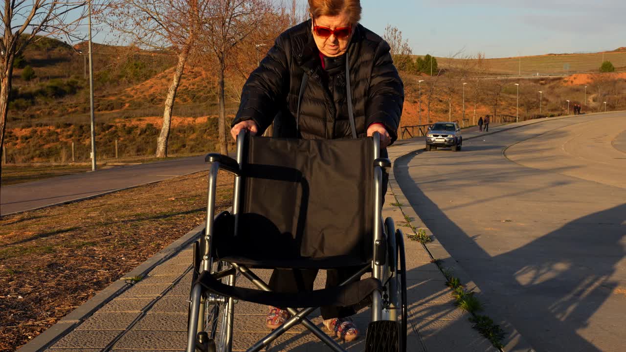Elderly woman walking up with an empty wheelchair on a sunny outdoor path