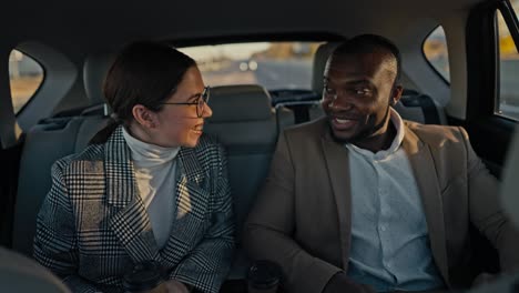 Confident-man-Businessman-with-Black-skin-color-in-a-brown-suit-talks-about-his-ideas-and-plans-to-his-colleague-a-brunette-girl-in-round-glasses-and-a-gray-jacket-during-his-trip-in-a-modern-car-outside-an-urban-area