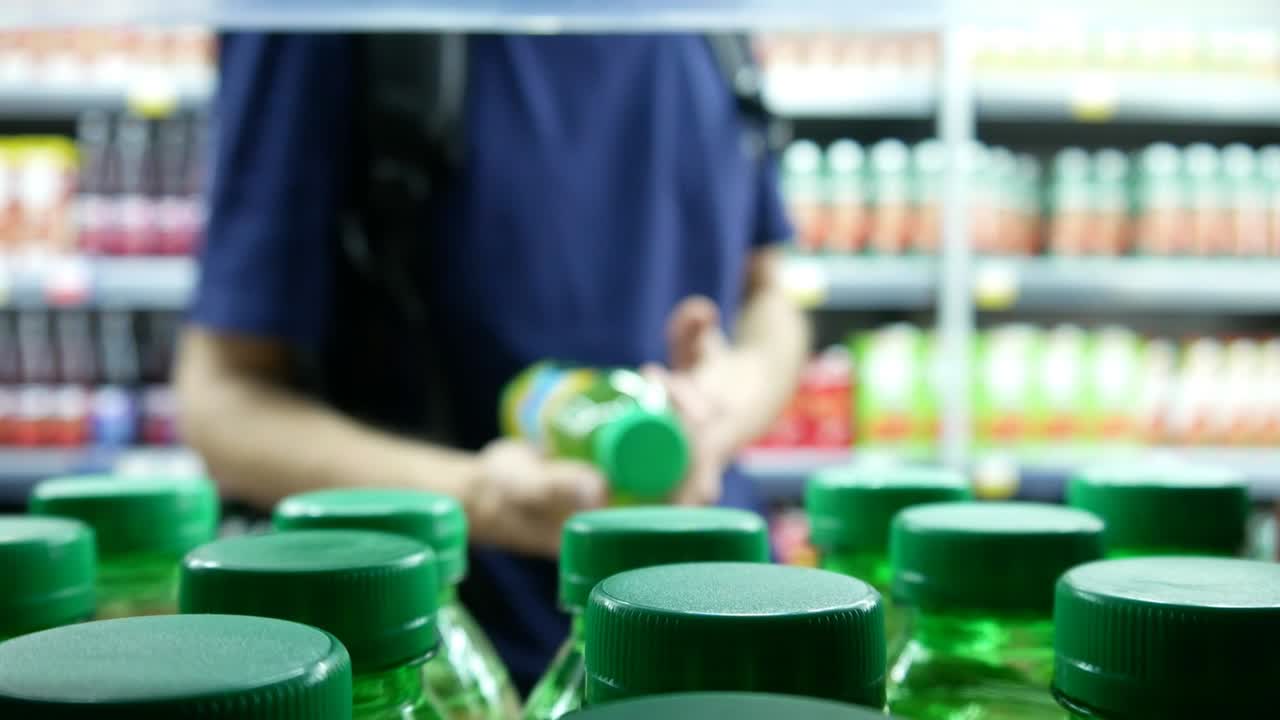 Close-up of many tea or juice bottles with green caps on a supermarket shelf and a young man takes one